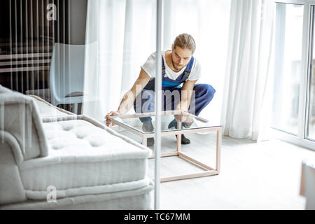Handsome mover in uniform fixing coffee table at the living room, delivering some furniture Stock Photo