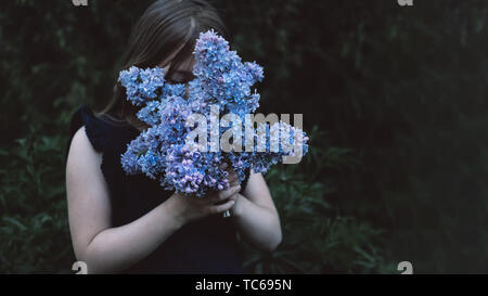 Cute little girl smelling lilac. Person holding flowers on dark natural background. Child enjoying bouquet outside. Going to park, forest in summer and spring. Connection with nature idea Stock Photo