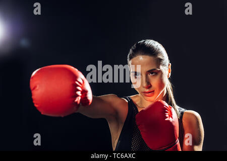Focused female boxer in red boxing gloves training and looking away on black Stock Photo