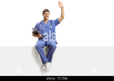 Full length portrait of a young male doctor in a blue uniform sitting on a white panel and greeting with hand isolated on white background Stock Photo