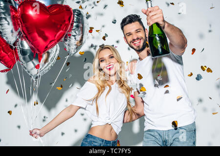 selective focus of cheerful bearded man holding bottle of champagne near woman with balloons near confetti on white Stock Photo