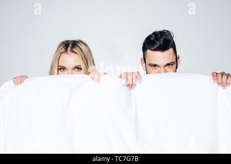 blonde woman and man looking at camera while covering face with blanket on white Stock Photo