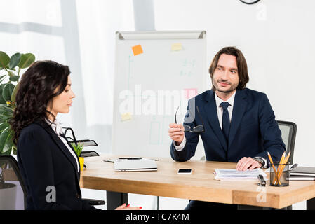 handsome recruiter holding glasses and looking at attractive woman in office Stock Photo