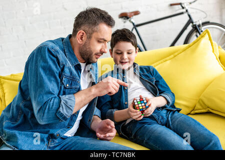 father sitting on couch with son and pointing with finger at toy cube Stock Photo