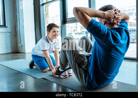 smiling son helping father sitting on fitness mat and doing sit up exercise at gym Stock Photo
