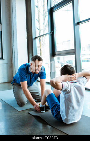 father helping son sitting on fitness mat and doing sit up exercise at gym Stock Photo