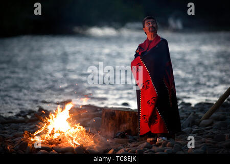 Mid-adult man with a blanket around him as he stands beside a bonfire on a rocky beach Stock Photo