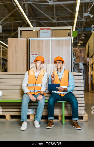 thoughtful multicultural workers in safety vests and helmets sitting on bench in warehouse Stock Photo