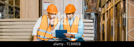 panoramic shot of serious multicultural workers sitting on bench in warehouse and looking at clipboard Stock Photo