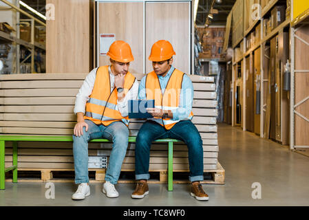concentrated multicultural workers sitting on bench in warehouse and looking at clipboard Stock Photo