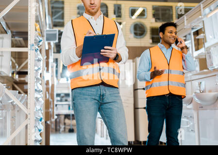 cropped view of worker writing in clipboard near indian colleague talking on smartphone Stock Photo