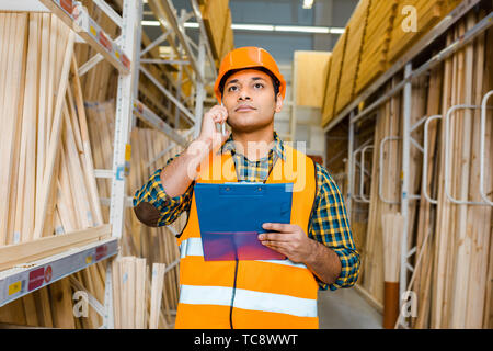 serious indian worker talking on smartphone and holding clipboard in warehouse Stock Photo