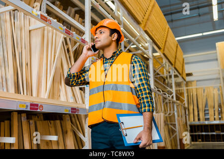handsome indian storehouse worker talking on smartphone and holding clipboard Stock Photo
