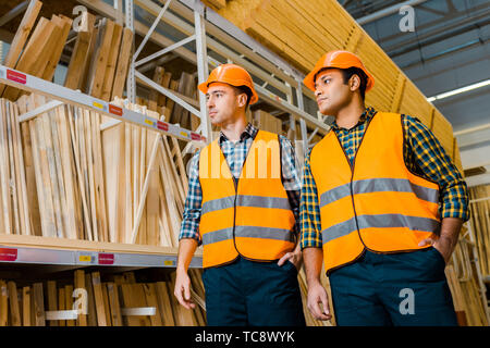 multicultural warehouse workers in safety vasts and helmets standing near racks with wooden construction materials Stock Photo