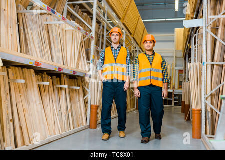 multicultural warehouse workers in safety vasts and helmets walking along racks with wooden construction materials Stock Photo