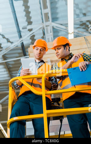 serious multicultural warehouse workers using digital tablet while standing on scissor lift Stock Photo