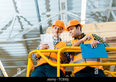 thoughtful multicultural workers using digital tablet while standing on scissor lift in warehouse Stock Photo