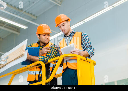 thoughtful multicultural workers with digital tablet and clipboard standing on scissor lift Stock Photo