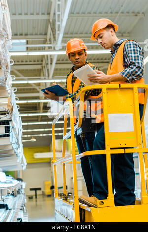 serious multicultural workers with digital tablet and clipboard standing on scissor lift in warehouse Stock Photo