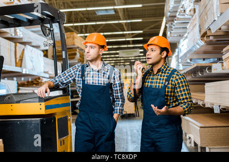 multicultural warehouse workers in uniform and helmets standing near forklift machine in storehouse Stock Photo