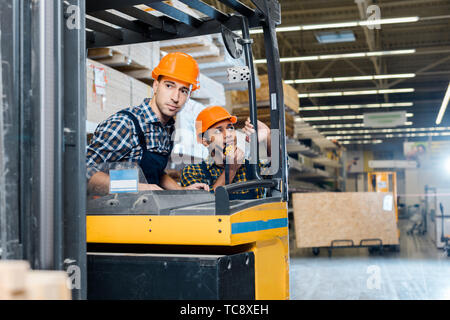 attentive worker sitting in forklift machine near his indian colleague talking on walkie talkie Stock Photo