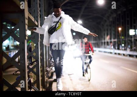 young man balancing on metal construction of bridge Hackerbrücke, Hacker bridge, at night in city, next to street, bicycle driver, in Munich, Germany