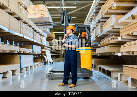 indian warehouse worker sitting in forklift machine near thoughtful colleague with clipboard Stock Photo
