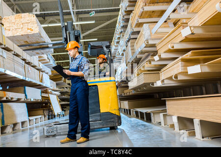 concentrated indian warehouse worker sitting in forklift machine near thoughtful colleague writing in clipboard Stock Photo