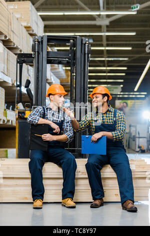 two smiling multicultural workers with clipboards talking while sitting on plywood Stock Photo