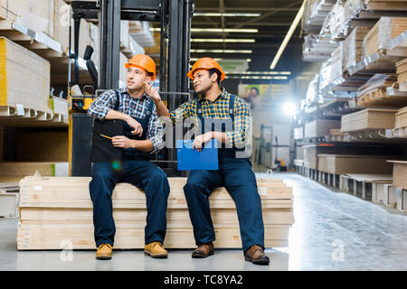 two multicultural workers talking while sitting on plywood in warehouse Stock Photo