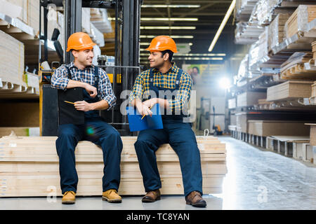 two multicultural workers smiling and talking while sitting on plywood in warehouse Stock Photo