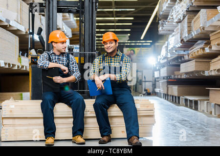 two smiling multicultural workers with clipboards sitting on plywood Stock Photo
