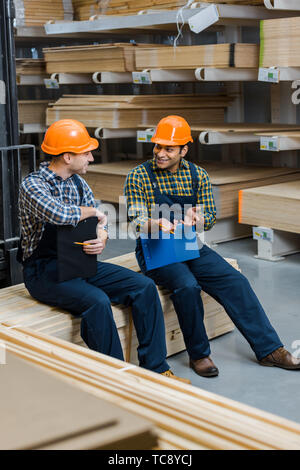 two smiling multicultural workers with clipboards talking while sitting in warehouse Stock Photo