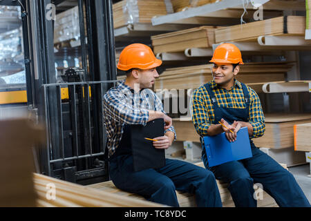 selective focus of two smiling multicultural workers sitting in warehouse Stock Photo