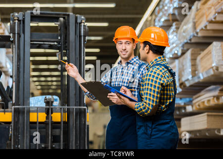 smiling warehouse worker in uniform and helmet talking to indian colleague Stock Photo