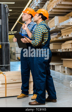 two multicultural workers in uniform holding clipboards while standing in warehouse Stock Photo