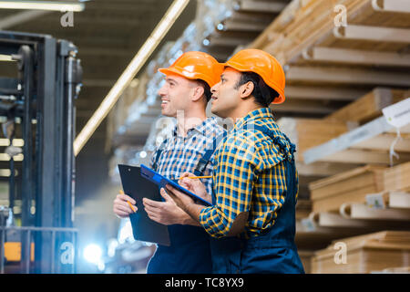 two multicultural warehouse workers smiling while holding clipboards Stock Photo