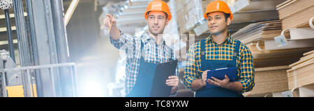 panoramic shot of warehouse worker pointing with finger while standing near indian friend Stock Photo