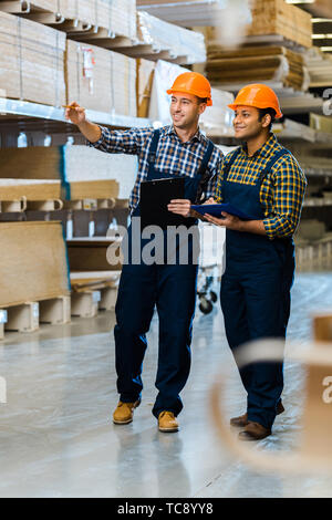 smiling warehouse worker in uniform pointing with finger near indian colleague Stock Photo