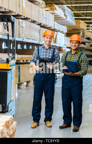two smiling multicultural workers in overalls and safety vasts smiling and looking at camera Stock Photo