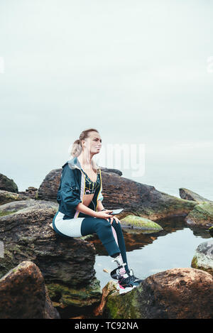 Enjoying nature and music. Happy disabled athlete woman in sportswear with prosthetic leg sitting on the stone and listening music. Sport concept. Music concept. Disabled Sportsman. Stock Photo