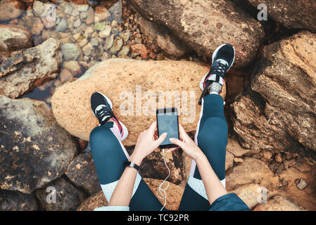 Top view of disabled woman in sport wear with leg prosthesis sitting on the boulders and holding smart phone. Digital concept. Workout. Disabled Sportsman. Healthy lifestyle. Stock Photo