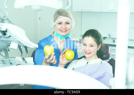 Happy teen girl sitting in dental chair with female dentist, showing fresh apples after  successful dental treatment Stock Photo