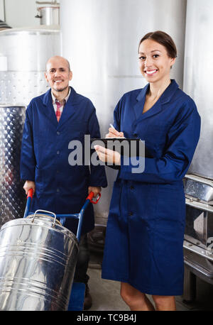 Glad diligent female winery worker taking notes while male one working in fermentation section Stock Photo