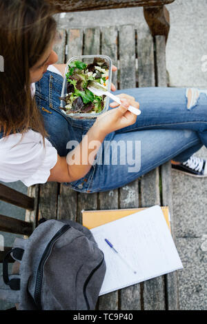 Midsection portrait of a student girl sitting in a bench while eating healthy salad with pasta in a glass lunch box Stock Photo