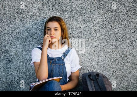 Pensive teen student girl in denim overalls studying and listening to music sitting on the floor Stock Photo