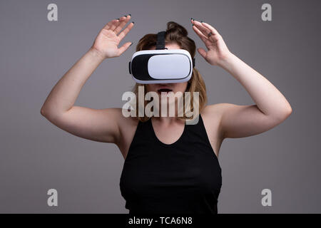 Female using VR gear is holding her hands up as displaying a shocked expression. Studio shot against gray background Stock Photo