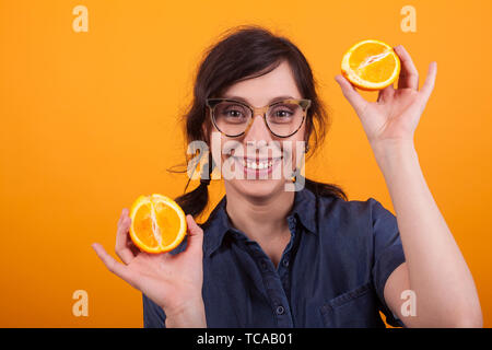 Portrait of attractive young woman with glasses smiling and showing fresh oranges at the camera in studio over yellow background. Cheerful young woman with summer citrus orange. Pretty girl holding jucy oranges . Stock Photo