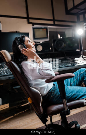 pretty, smiling mixed raced sound producer listening music in headphones near mixing console Stock Photo