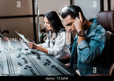 selective focus of handsome mixed race sound producer in earphones near pretty colleague working at mixing console Stock Photo